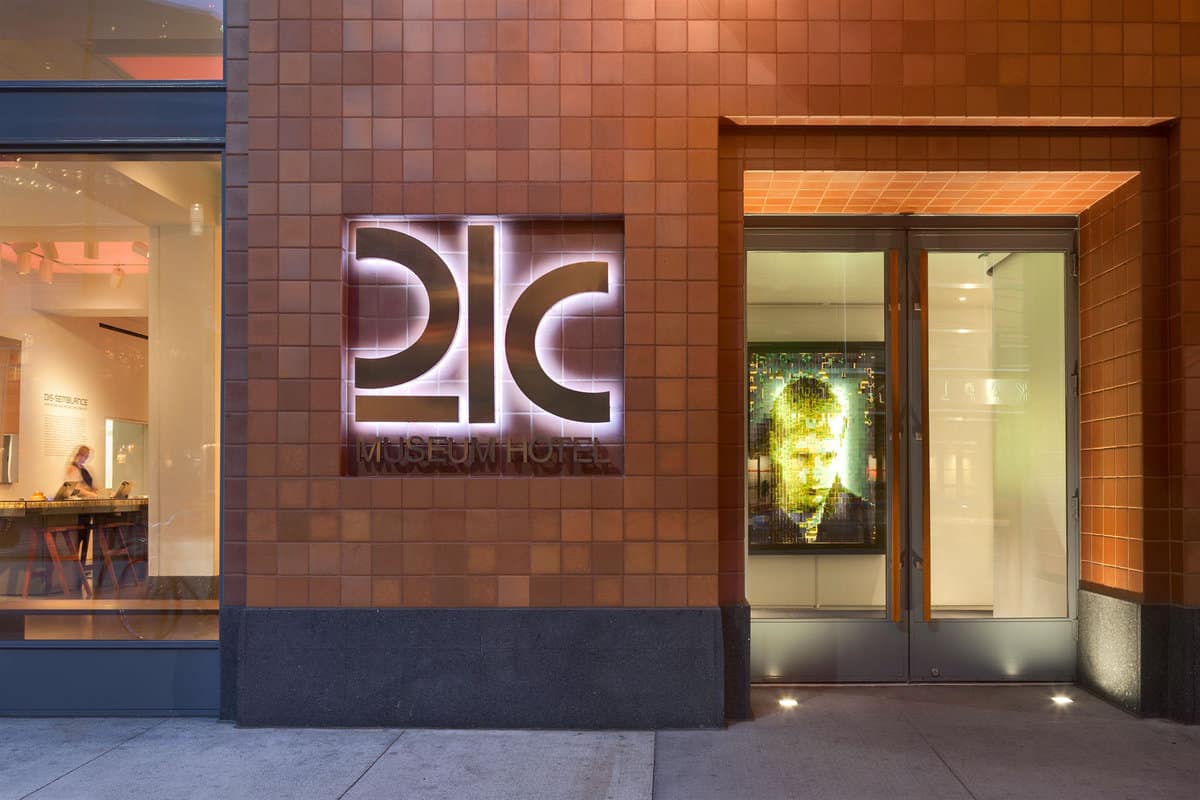 Exterior view of the 21c Museum Hotel entrance at dusk, featuring illuminated signage on a terra cotta tile facade, with visible interior dining area and artwork.
