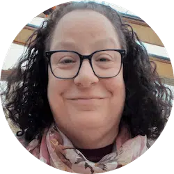 A person with shoulder-length curly dark hair and wearing black-rimmed glasses smiles at the camera. They are dressed in a dark top and a floral scarf, positioned indoors with beams and windows in the background.