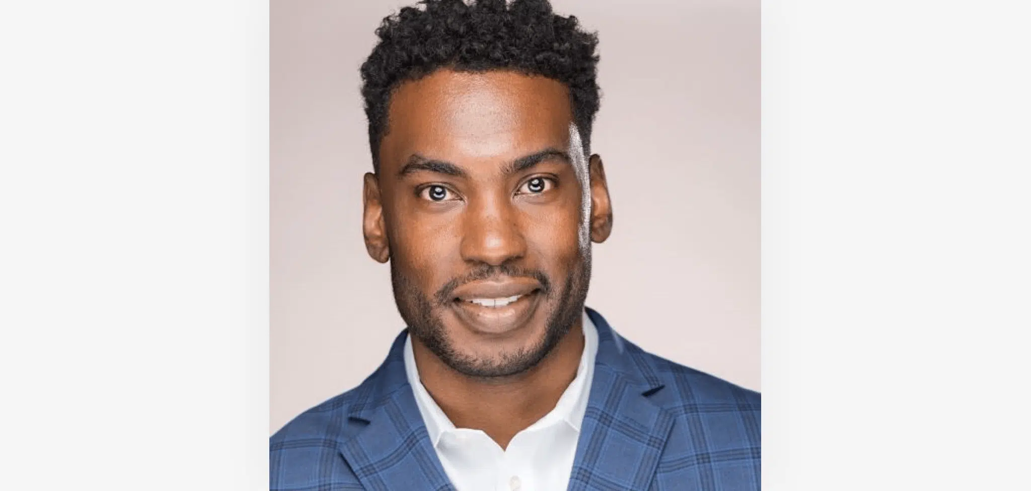 A man with short, curly black hair and a neatly trimmed beard smiles at the camera. He is wearing a blue checkered blazer over a white dress shirt. The background is a soft, light color, making him stand out prominently in the image.