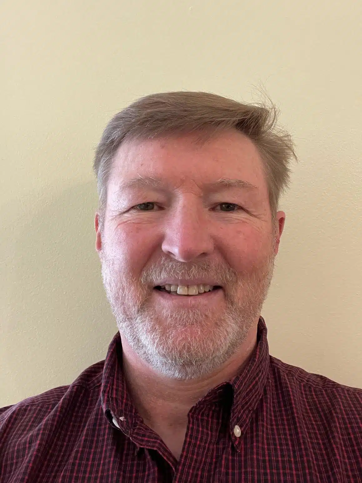 A man with short hair, a beard, and a mustache is smiling at the camera. He is wearing a maroon button-up shirt and standing against a plain beige background.