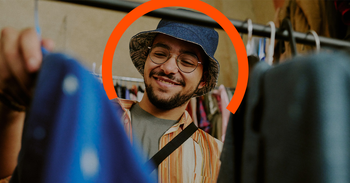 A man wearing a blue bucket hat and glasses smiles while browsing clothes on a rack, with an orange semi-circle graphic behind his head—showcasing what retail employers need to know about engaging shoppers.