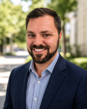 A man with short dark hair and a beard, wearing a blue suit jacket and light blue shirt, smiles outdoors with trees and a building in the background.