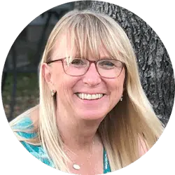 A woman with long blonde hair, wearing glasses and a teal top, is smiling outdoors. She has a pendant necklace and small earrings, with a tree trunk visible in the background.