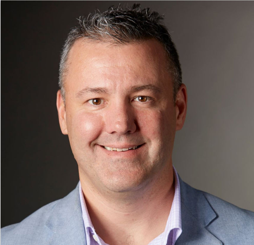 A man with short gray hair and a light beard, wearing a light blue blazer and light purple shirt, smiling at the camera against a plain background.
