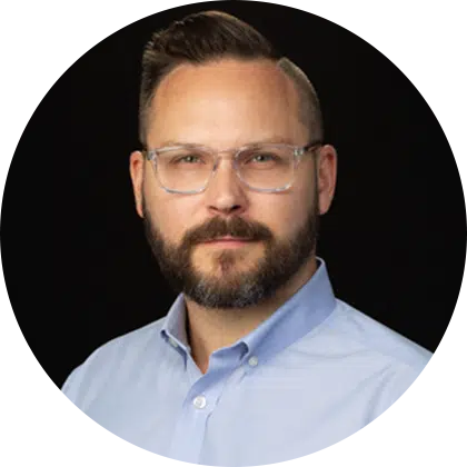 Portrait of a man with a beard, wearing glasses and a light blue button-up shirt. He has short, styled hair and a neutral expression, set against a black background.