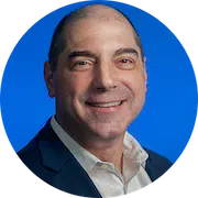 A bald man with short black hair on the sides, wearing a dark blue suit and a white collared shirt, is smiling. The background is a solid, vibrant blue. The image is cropped in a circular frame.
