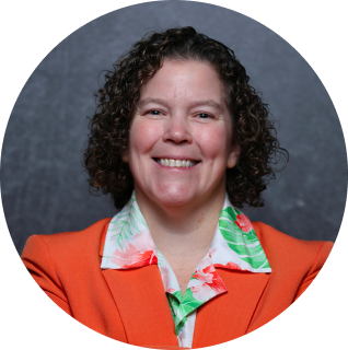 A person with short curly hair, wearing an orange blazer and a floral-patterned shirt, smiles in front of a dark gray background.