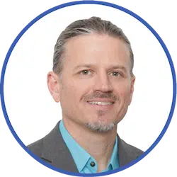A man with greying hair slicked back, a short goatee, and a friendly smile is wearing a light blue collared shirt and a grey blazer. The photo is cropped to a circular frame with a blue border against a white background.