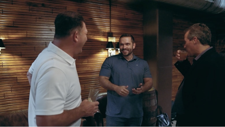 Three men stand in a room with wooden walls, engaged in conversation. One man in a white shirt laughs, a man in a blue shirt smiles, and a man in a black shirt takes a sip from a glass.