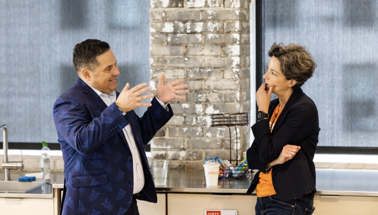 Two people are engaged in a conversation in a modern kitchen area with large windows, exposed brick walls, and a countertop with various items, including chips and a sanitizer bottle.