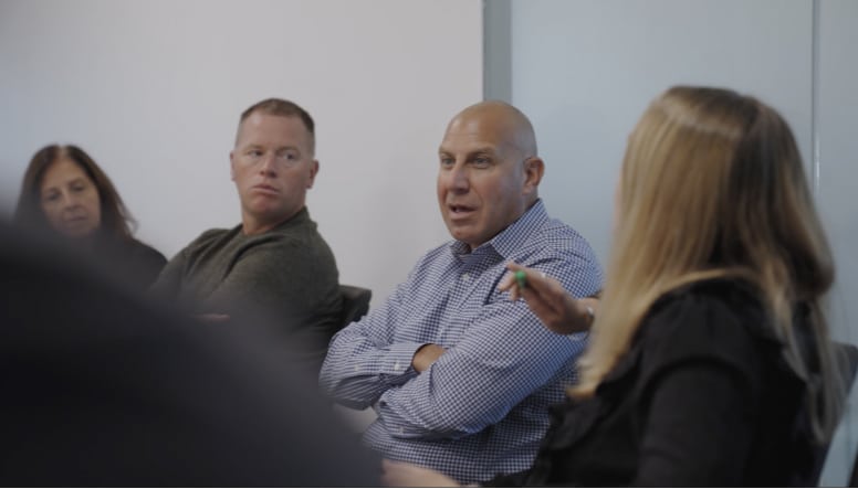 A group of four people seated in a meeting room, engaged in a discussion. One person is speaking while others listen attentively.