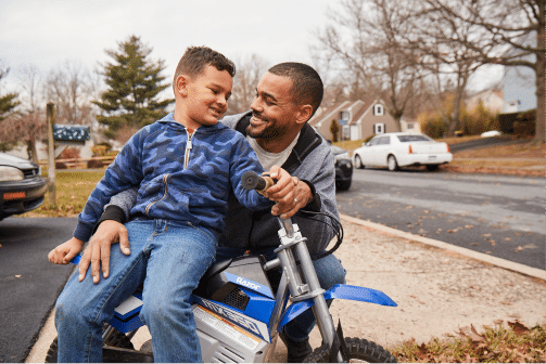 A man and a boy sit together on a small blue motorbike in a residential neighborhood. Both are smiling and looking at each other. Trees and parked cars are visible in the background.