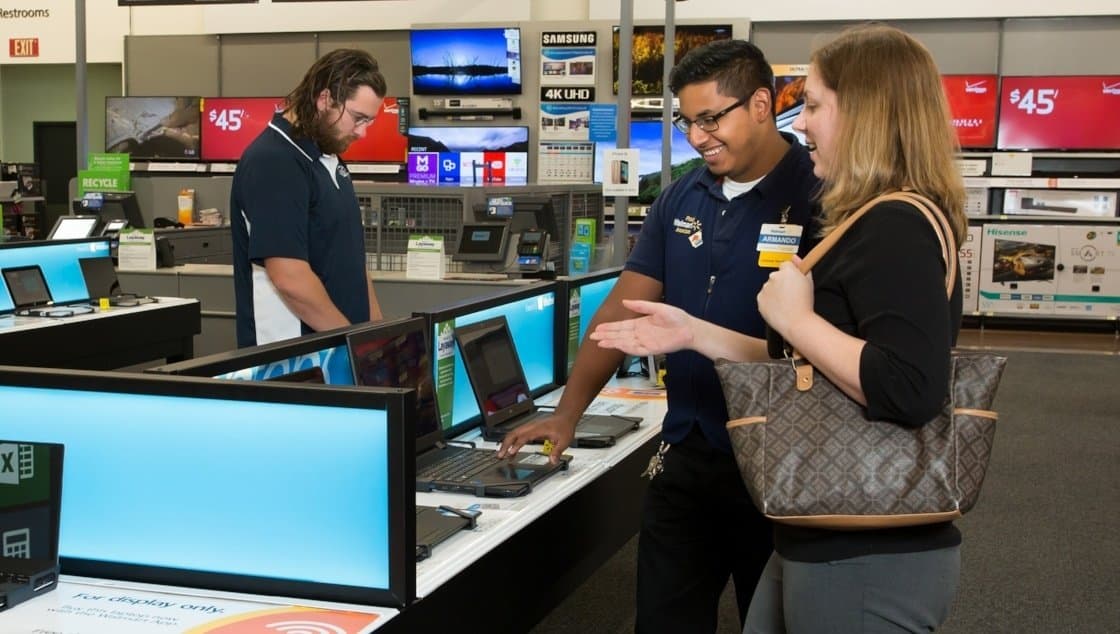 Two store employees are assisting a female customer in an electronics store. One employee is demonstrating a laptop, while the other is working at a counter. The woman is smiling and engaging with the employee. Various electronics and displays are visible in the background.