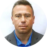 A man with short brown hair wearing a blue shirt and gray blazer is looking directly at the camera in this headshot photo.