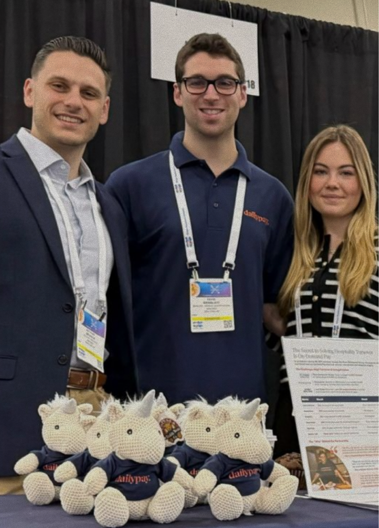 Three people stand behind a table displaying crocheted unicorn toys and an informational flyer at a conference booth.
