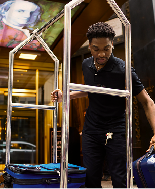 A hotel staff member wearing a black shirt loads blue suitcases onto a luggage cart outside a building entrance.