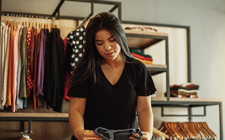 A woman with long dark hair wearing a black shirt looks down at an item of clothing in a store with racks of clothes behind her.