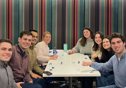 Eight people sit around a white table, smiling at the camera. The table has small objects on it, and the background features a colorful striped wall.