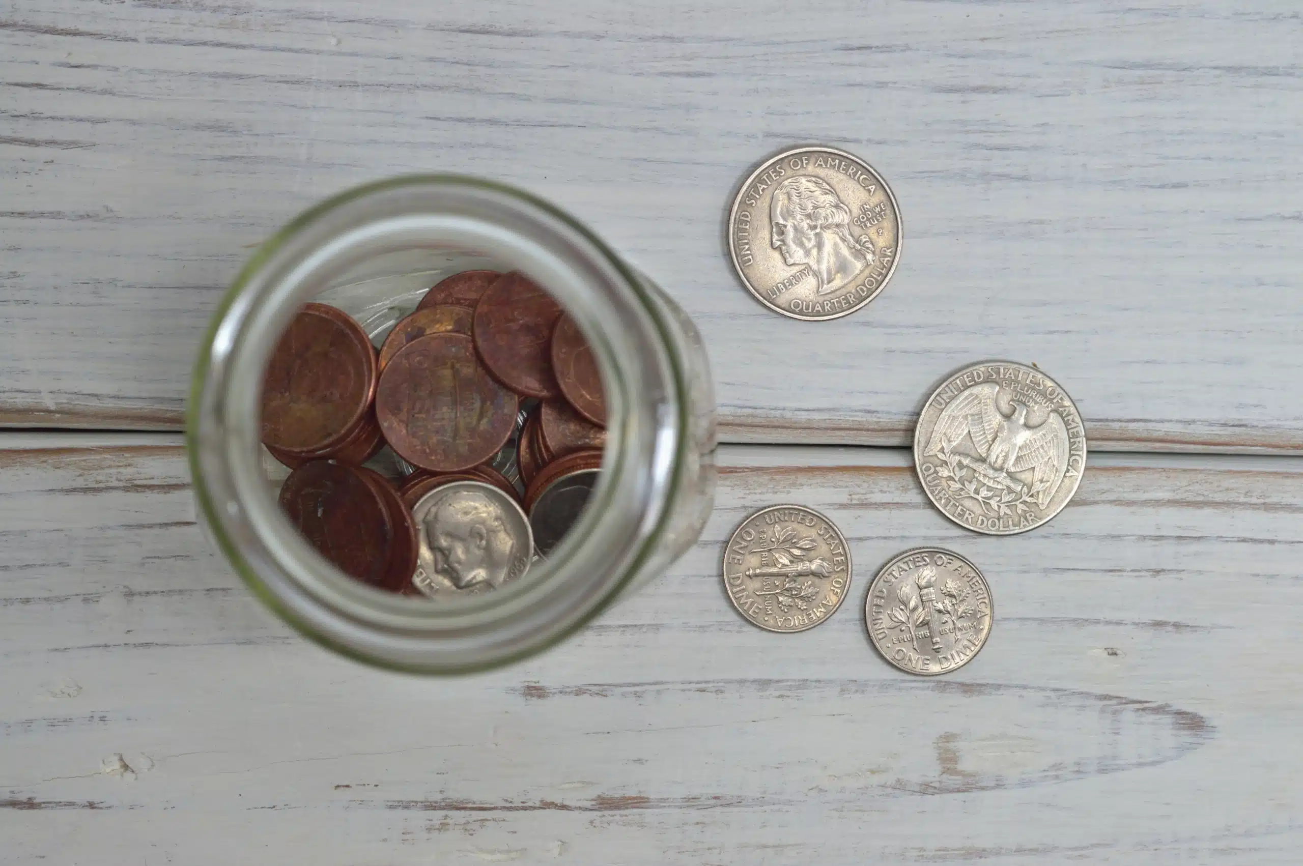 A jar filled with pennies viewed from above, placed on a wooden surface. Nearby, there are several quarters and dimes scattered on the table.