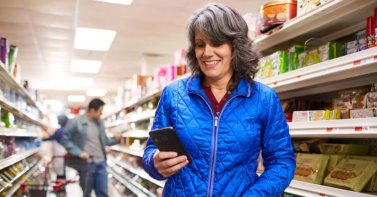 A person wearing a blue jacket is smiling and looking at their phone in a grocery store aisle, possibly checking their Ceridian Dayforce Wallet. Shelves stocked with various products are visible in the background.