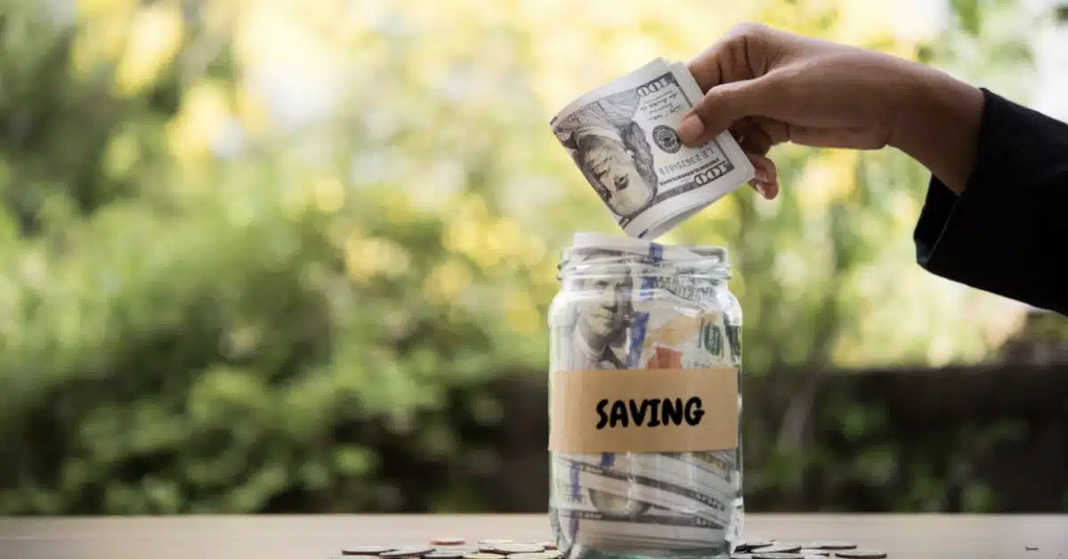 A hand places U.S. dollar bills into a jar labeled "Saving," with coins scattered around the jar on a wooden surface in the foreground.