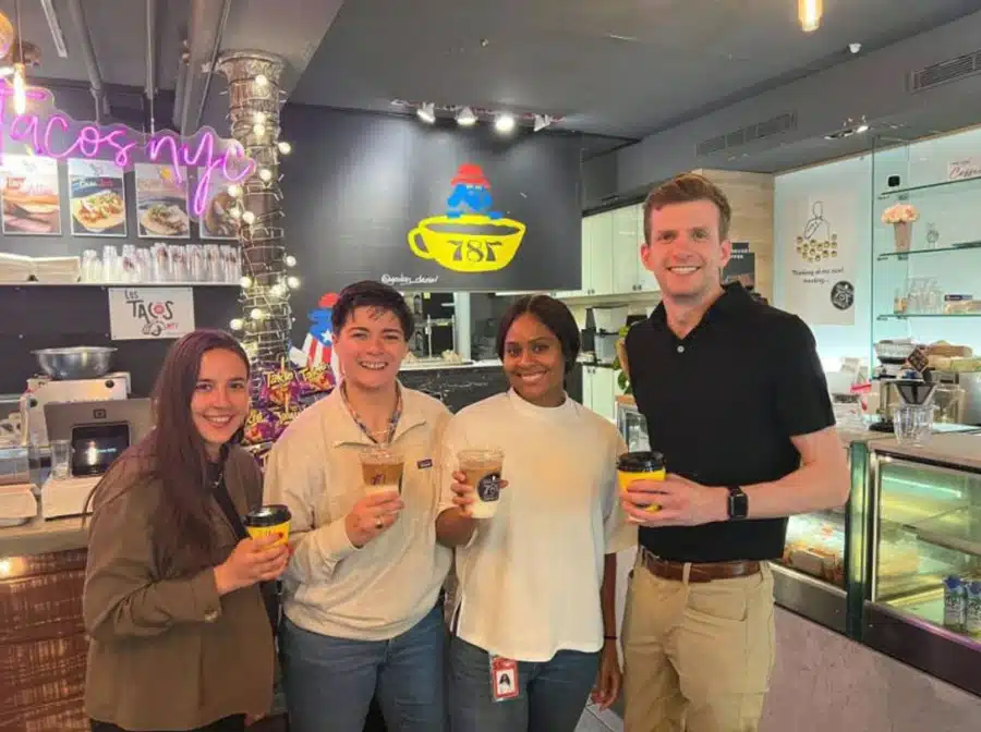 Four people smiling and holding drinks, standing inside a cafe with a colorful neon sign and a counter displaying pastries in the background.