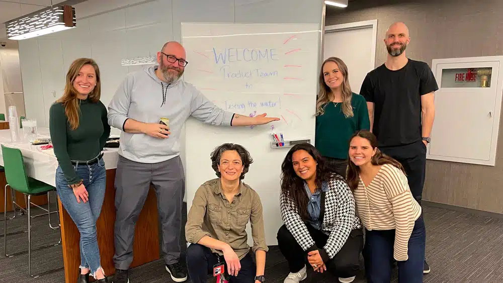 A group of seven people pose in front of a whiteboard that reads "WELCOME Product Team to 'Testing the water'." They are smiling and wearing casual clothes in an office break room.