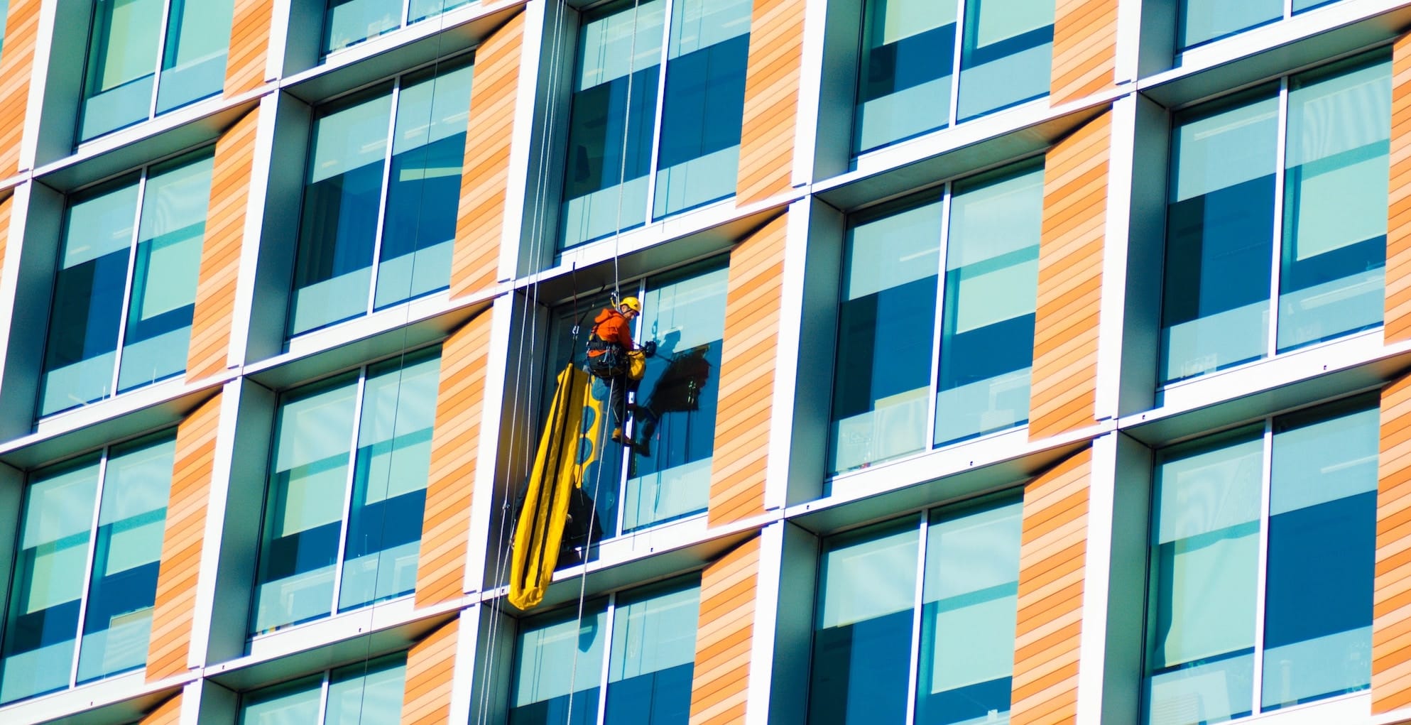 A window cleaner in safety gear is suspended on a platform cleaning a multicolored, geometric-patterned high-rise building under a clear blue sky.
