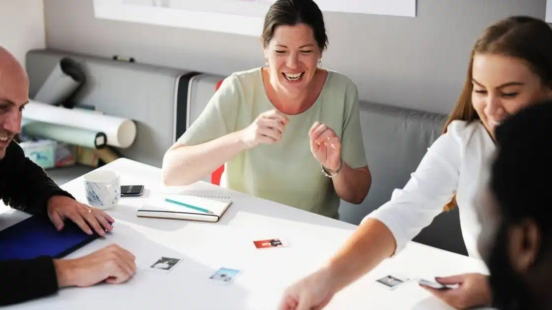 Four people sitting around a table, engaged in a lively discussion, with papers and photos spread out on the table. One woman is smiling and gesturing, while another person reaches for a photo.