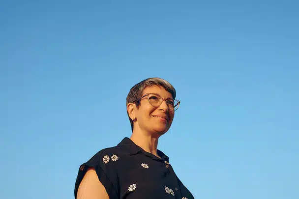 A person with short, graying hair and glasses smiles while looking slightly upward against a clear blue sky. They are wearing a black button-up shirt with small white floral patterns. The sunlight illuminates their face, giving a warm and cheerful expression.