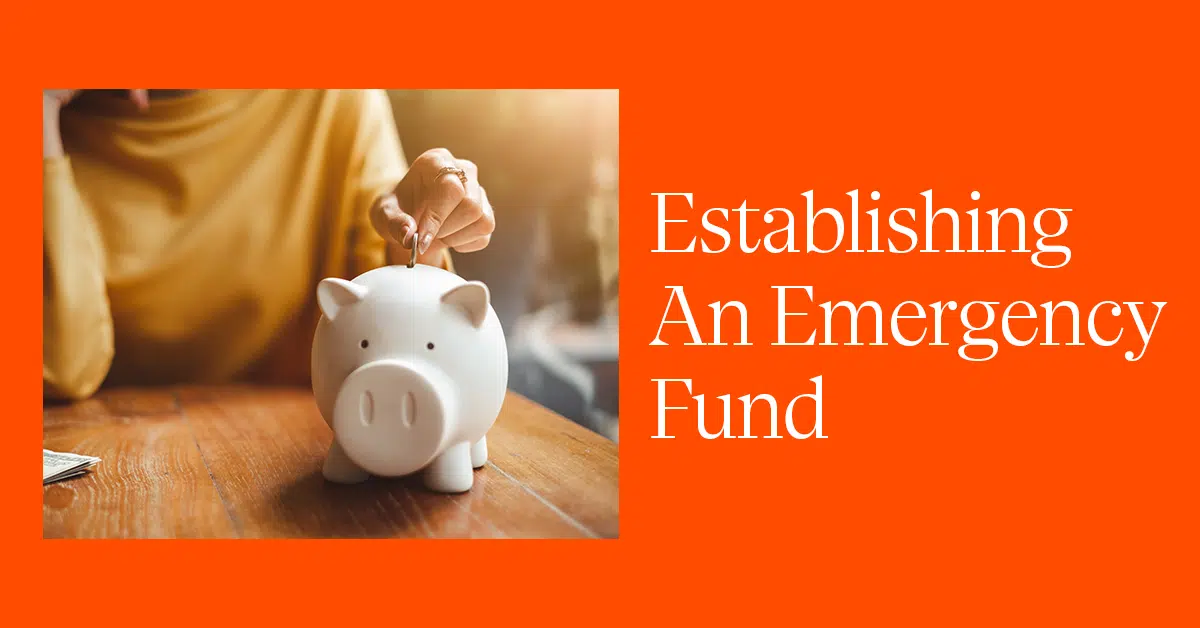 A person wearing a yellow sweater is placing a coin into a white piggy bank on a wooden table. The background is orange, and the text to the right of the image reads "Establishing An Emergency Fund" in white capital letters. Sunlight shines warmly onto the scene.