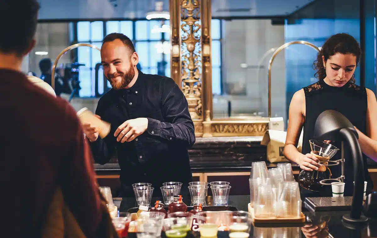 Two baristas, a man and a woman, are preparing drinks behind a counter in a café. The man is shaking a cocktail shaker while the woman is focused on a coffee machine.