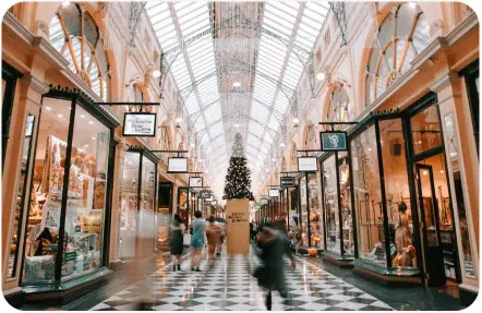 A spacious shopping mall with glass roof, arched ceilings, and illuminated storefronts on both sides. A decorated Christmas tree stands at the center, and people are walking by.