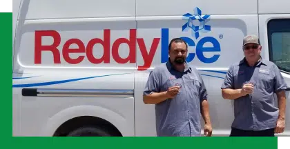 Two men in grey uniforms stand in front of a Reddy Ice delivery truck, each holding a small bag of ice.