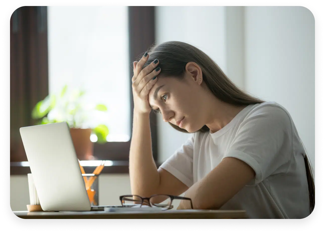 A woman with long hair is sitting at a desk with her hand on her forehead, looking at a laptop screen. Glasses and a cup with pens are on the desk. There's a plant in the background.