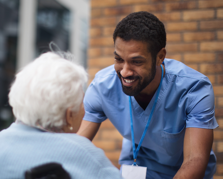 A male nurse in blue scrubs smiles while talking to an elderly woman with white hair seated in front of him.