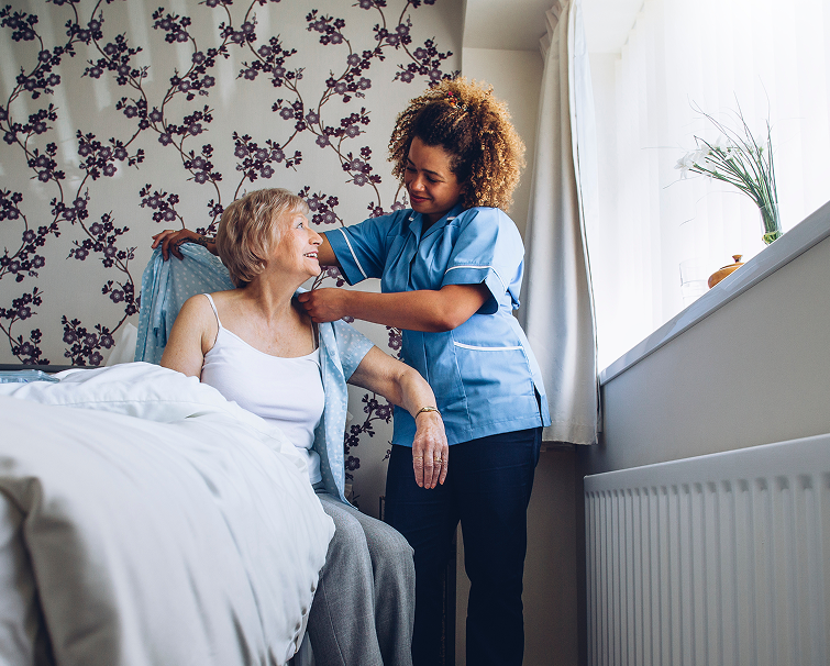 A caregiver in a blue uniform helps an older woman in a bedroom, adjusting her clothing as she sits on the edge of the bed.