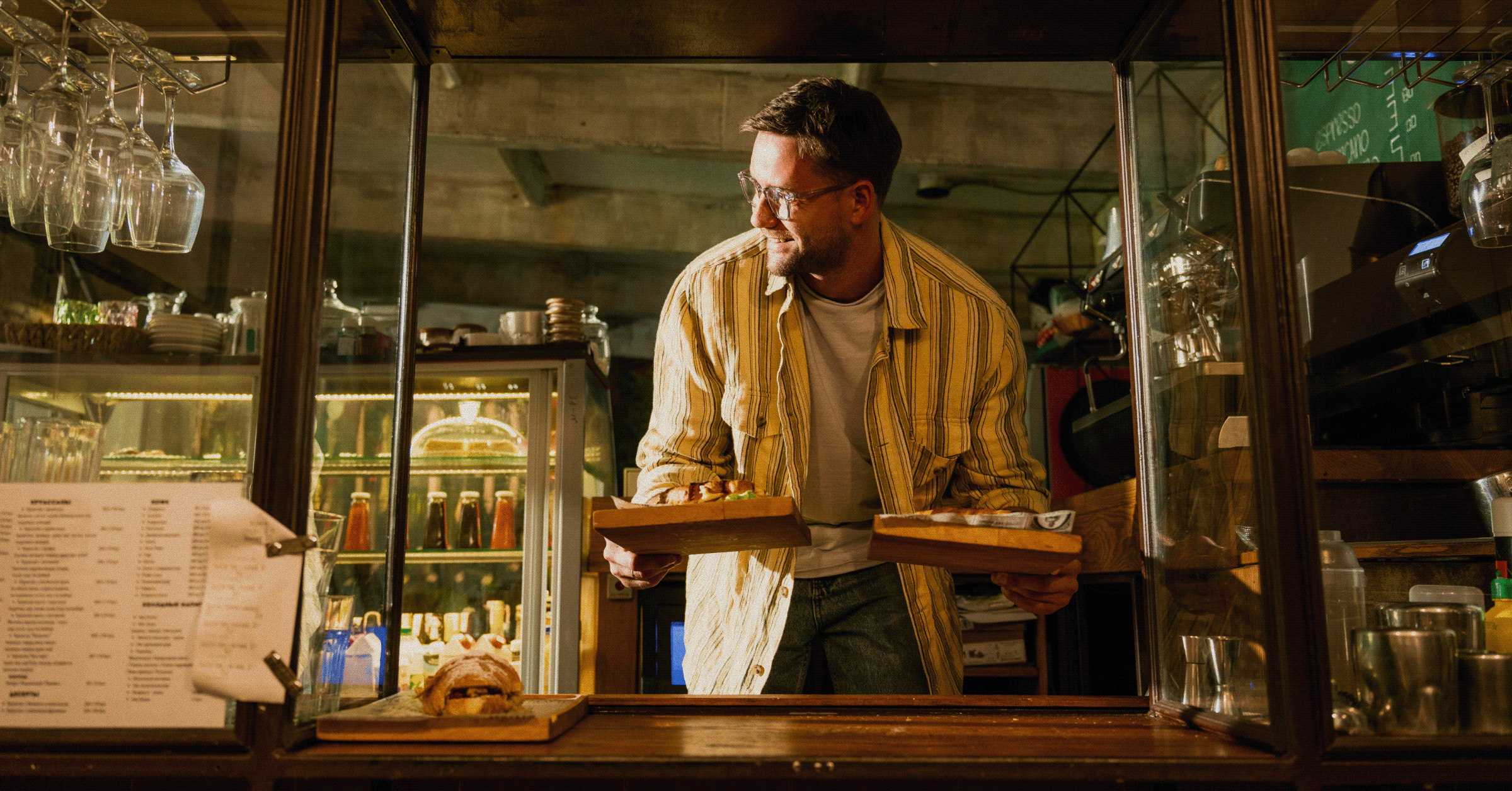 A barista in a yellow striped shirt reaching over a counter to serve two wooden trays of food.