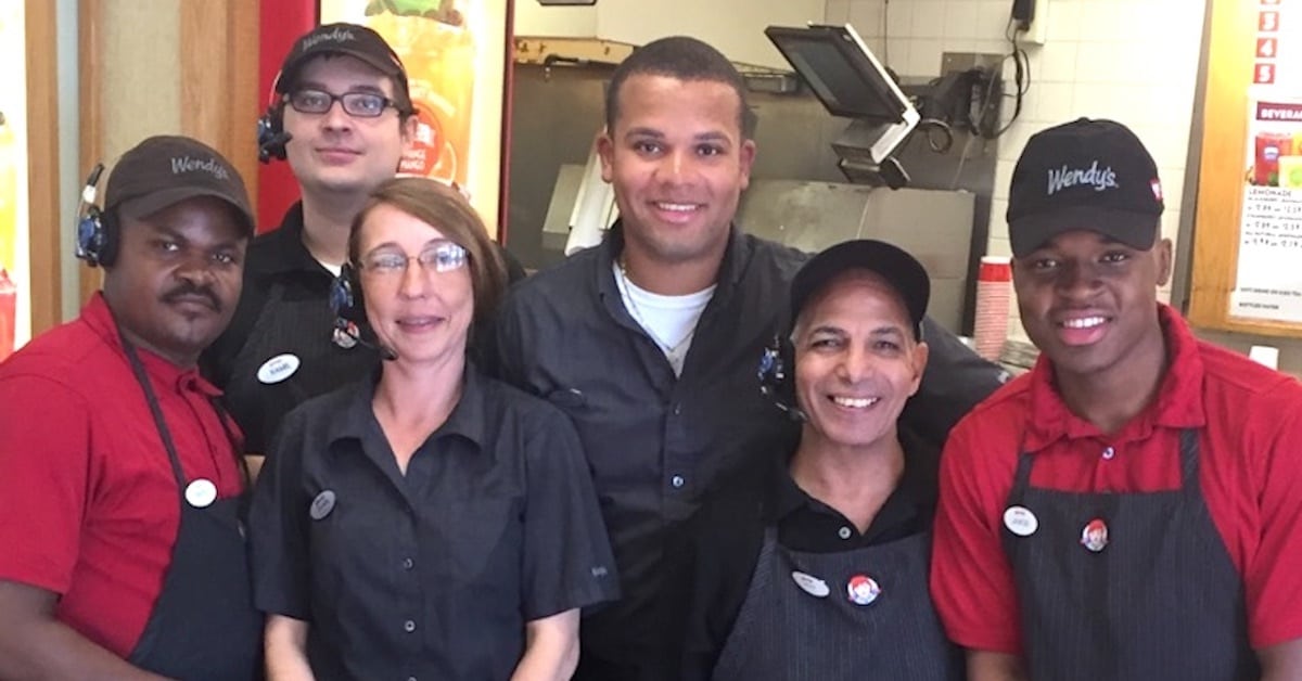 A group of six fast-food restaurant employees wearing Wendy's uniforms pose together inside the restaurant. Three employees are in red shirts, and three are in black shirts. They all stand close to each other, smiling at the camera, with kitchen equipment visible in the background.