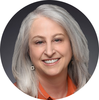 Portrait of a smiling woman with long, silver hair, wearing an orange top and square earrings, against a dark background.