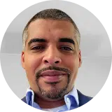 Close-up of a man with short hair and a goatee, wearing a blue collared shirt and a dark jacket. He is smiling slightly and looking directly at the camera with a neutral background.