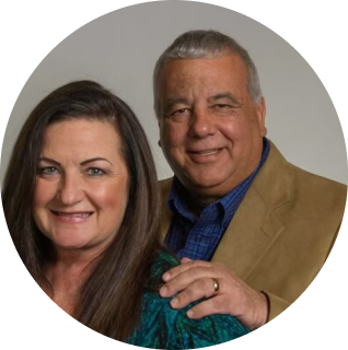A middle-aged woman with long brown hair and a man in a tan jacket and blue shirt pose together, smiling at the camera.