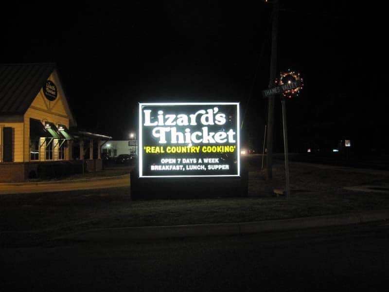 A brightly lit roadside sign at night displaying "Lizard's Thicket, Real Country Cooking," with information on open days and meal offerings, next to a dimly lit building and a street.