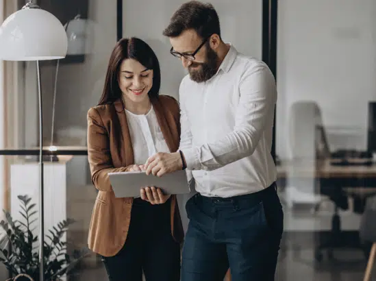 Two people, a woman and a man, stand in an office looking at a tablet together. They appear to be discussing something on the screen, perhaps analyzing data from the EWA Mercator 2021 report.