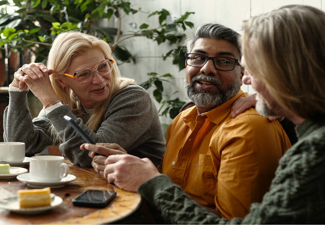 Three people sit at a table with coffee and cake, talking and smiling. One person holds a phone while the others listen. Indoor setting with plants in the background.