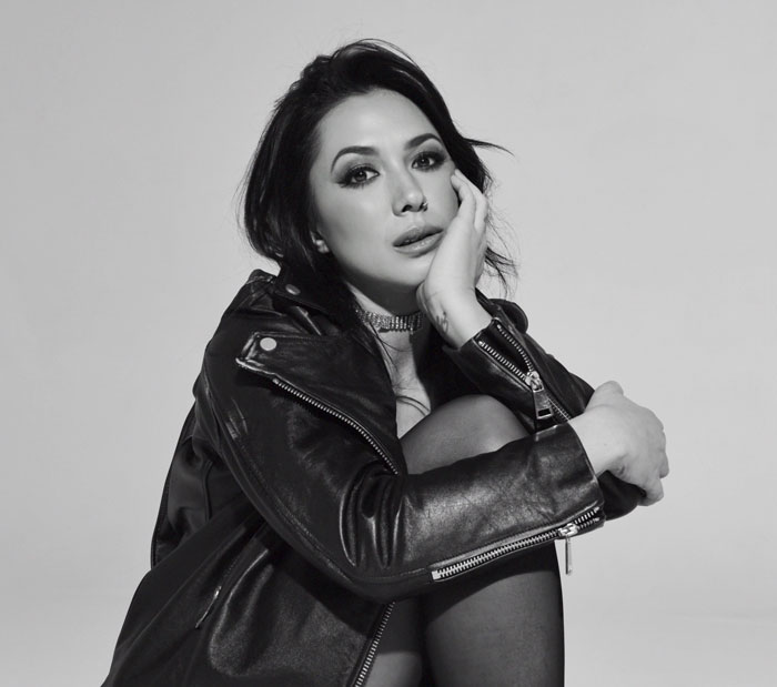 A woman wearing a leather jacket sits with one knee up, resting her head on her hand, looking at the camera in a black-and-white photo.