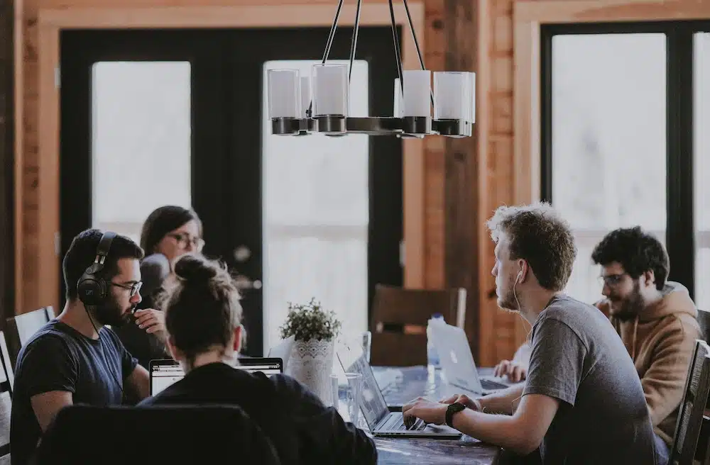 A group of people are working at a table with laptops in a room with wooden walls and large windows. A modern chandelier hangs above them.