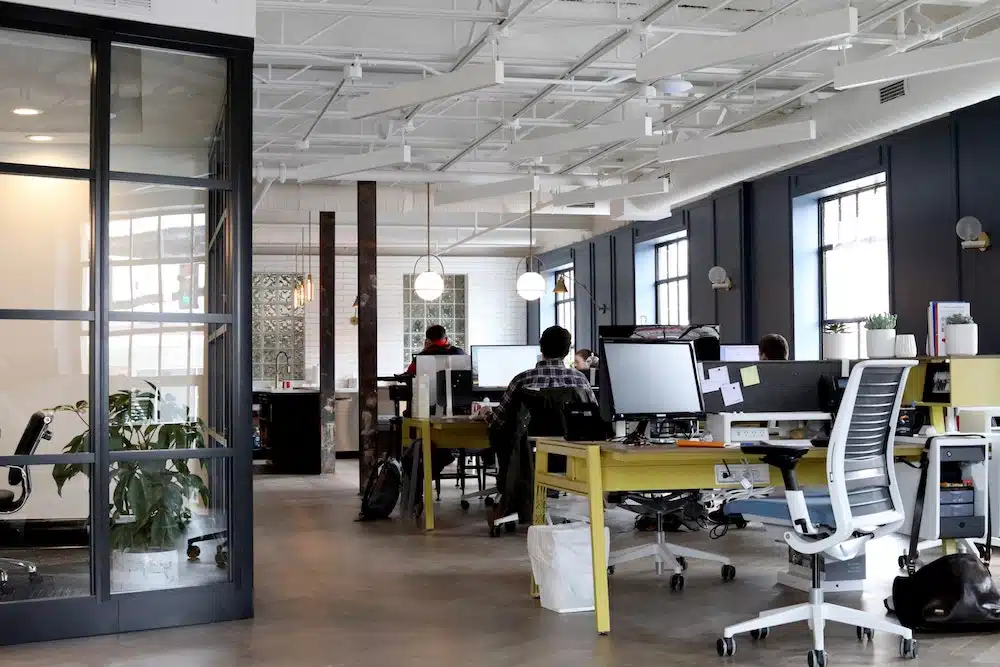 An open-plan office with several desks and chairs, computers, and people working. A glass-walled room and potted plants are visible in the background.