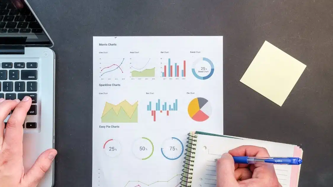 A person types on a laptop with their left hand and writes in a notebook with their right. A sheet with various colorful charts and a yellow sticky note are on the table.