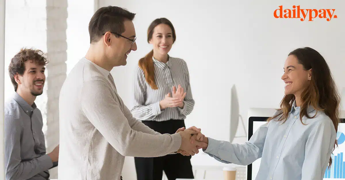 Two colleagues shaking hands in a bright office while two others, seated and standing, look on smiling. The word "dailypay" is displayed in the top right corner.